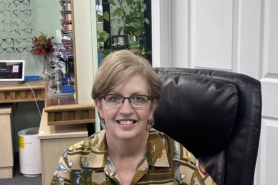 Rosemary, licensed optician, smiling at her desk in Rosemary's Optical Shop in Brockville with frames on the wall behind her
